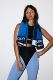 Woman wearing a blue towel with 'Rafale' branding, black top, and blue leggings on a white background.