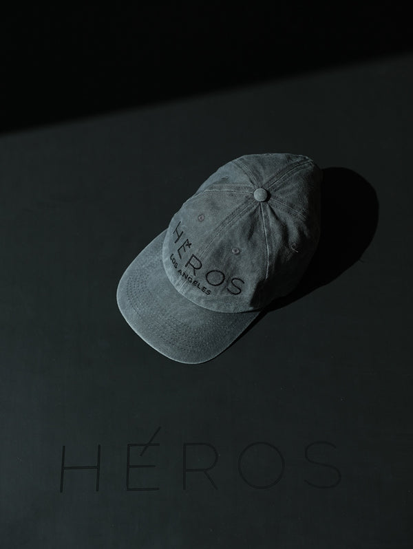 A washed black baseball cap with the HÉROS logo embroidered on the centre front, displayed on a dark background with the word 'HÉROS' also printed beside the cap.