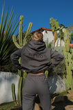 A person standing with their back towards the camera wearing a slate grey hooded sweatshirt with 'Héros' and 'Los Angeles' embroidered on it, paired with matching grey sweatpants, against a backdrop of cacti.
