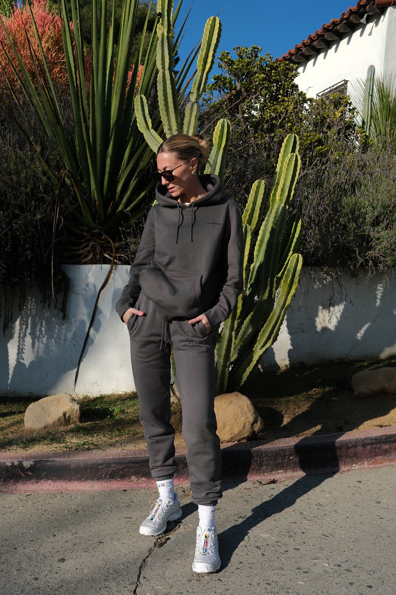 Person wearing a gray hoodie and pants standing in front of cacti and a white building.