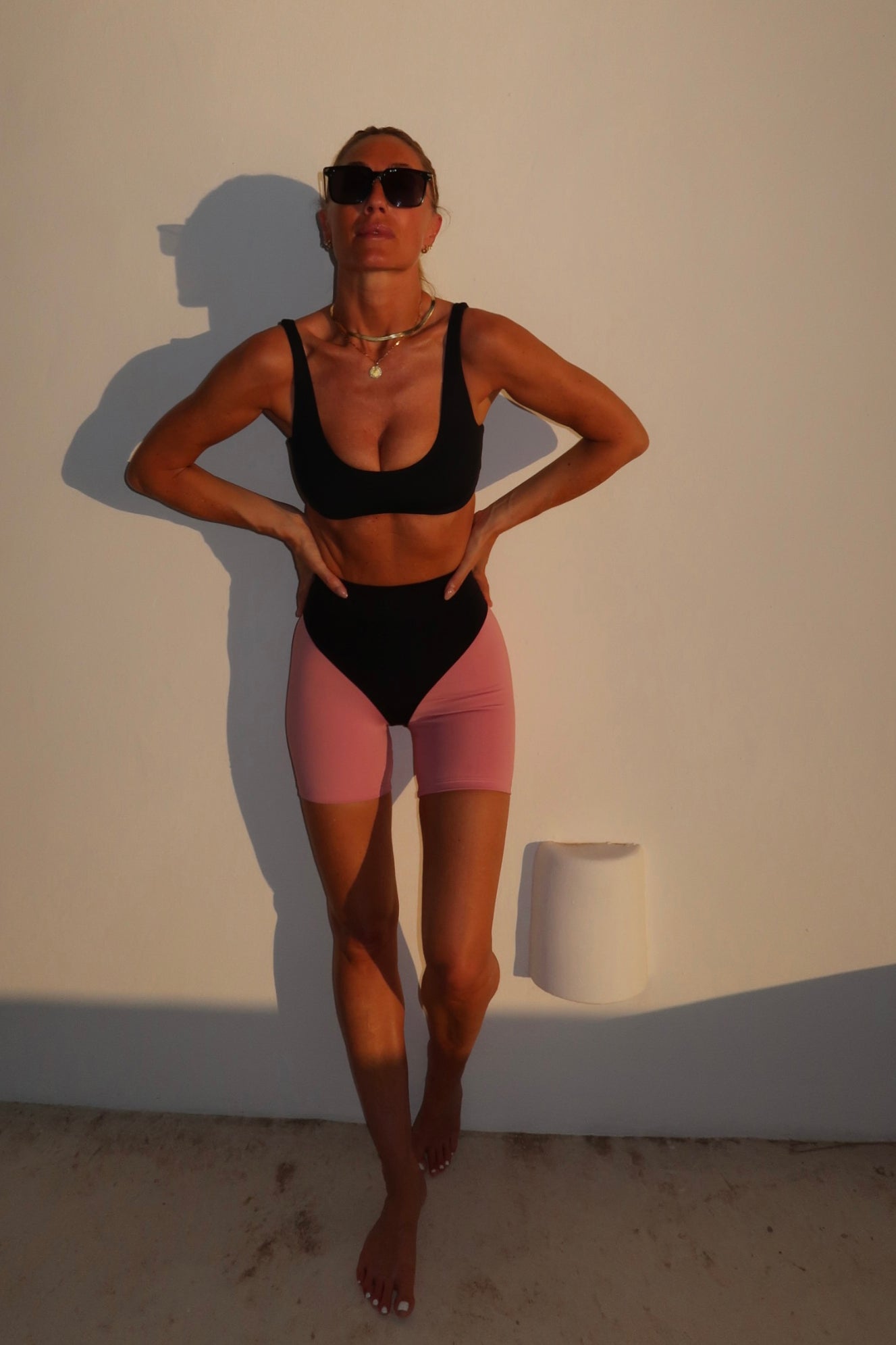 A woman modeling a two-piece swimsuit consisting of a black sports bra and matching black high-waisted briefs with pink leg portions, posing against a warm-lit beige wall.