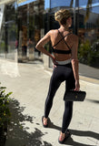 A woman wearing a black strappy top with a flattering curve neckline and thin straps, suitable for low-intensity workouts. She is also wearing black leggings and carrying a small black purse.
