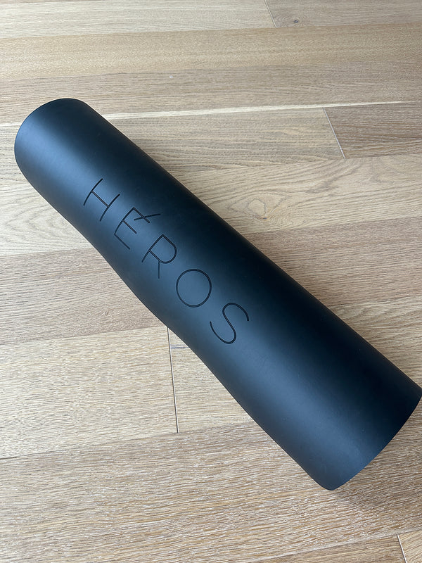 An oversized matte black yoga mat with the brand name 'HÉROS' engraved on it, displayed on a wooden floor.