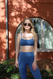 A woman standing outdoors wearing a blue sports bra and matching leggings. She has blonde hair and is wearing sunglasses.