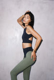 A woman posing in a black crop top with a keyhole front and cross back design, paired with green leggings.
