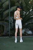 A person wearing a white unitard with a scoop neck and strap details on the back, standing on a tennis court.