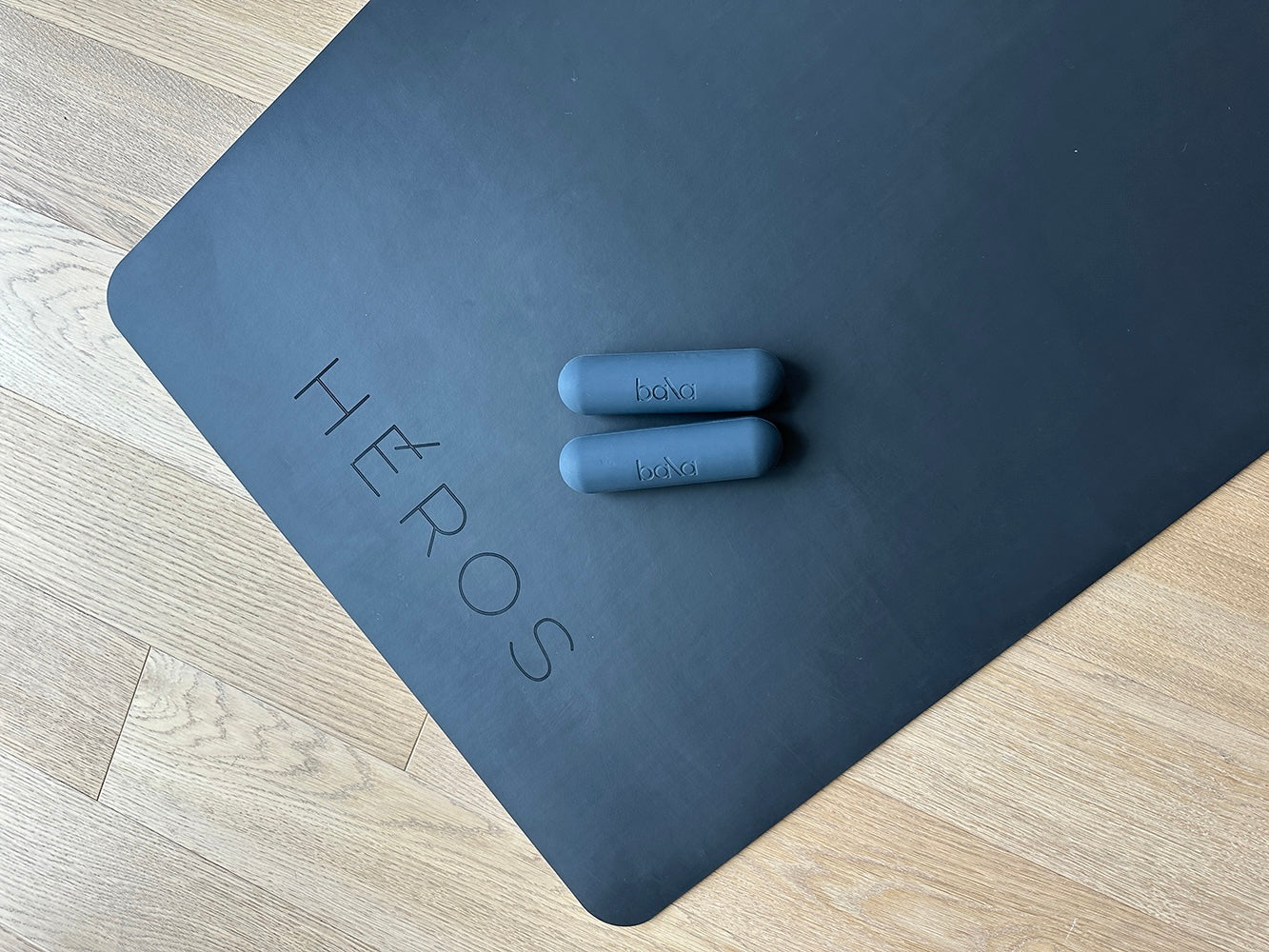 HÉROS black yoga mat with embossed logo and Bala hand weights on wooden floor — minimalist luxury fitness accessory setup.