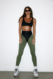 Woman wearing a black sports bra and green leggings with white sneakers on a white background