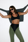 Woman in black sports bra and green leggings posing with arms outstretched against a white background