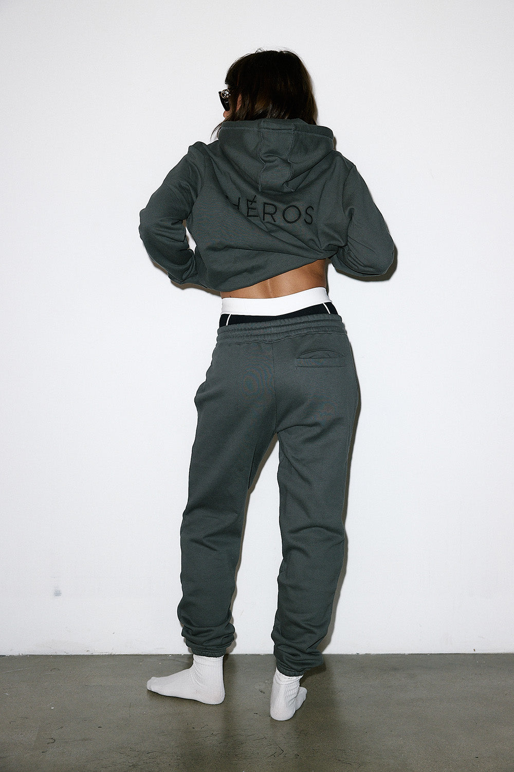 Person wearing a dark green tracksuit with 'HEROS' branding, standing against a white wall.