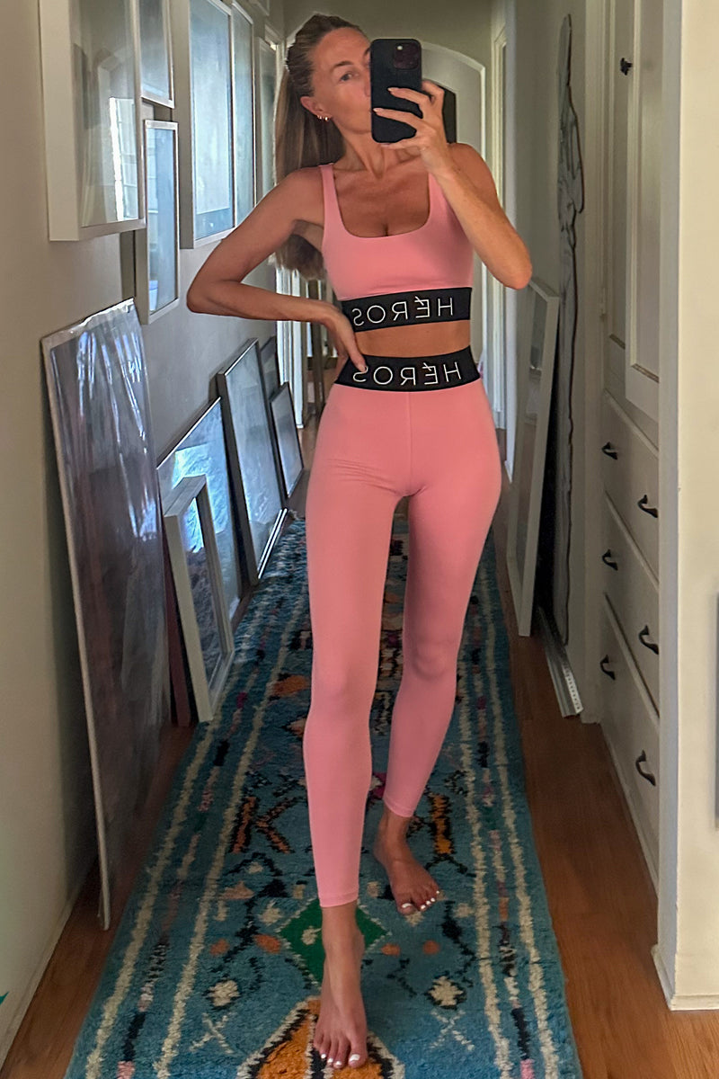 A woman takes a mirror selfie wearing a pink activewear set consisting of a sports bra and leggings with a black waistband that has white text. She is standing in a hallway with framed art and cabinets.