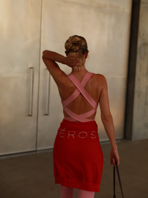 A woman shown from behind wearing a red mini skirt with 'HEROS' printed in white across the back and a pink strappy open-back top, standing indoors against a light concrete wall.